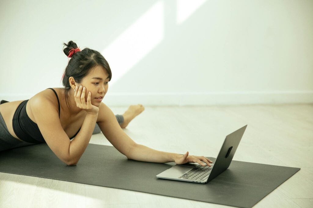 Asian woman practicing yoga at home, using a laptop. Bright daylight enhances focus and relaxation.